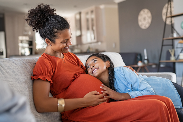 Happy mixed race daughter hugging belly of her expecting mother while relaxing on couch at home. African girl listening to baby movements while embracing pregnant woman. Pregnant black mom and future sister relaxing together on sofa at home.