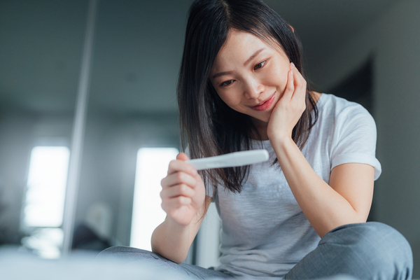 Young Asian woman holding home pregnancy test and looking cheerful. The longest wait ever. Life changing moment. Am I ready to be pregnant? Dream came true.
